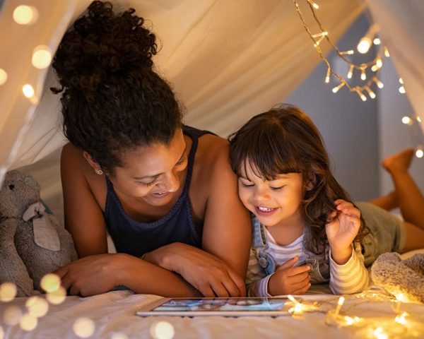 African mother and cute smiling girl using digital tablet while lying in illuminted tent in kid bedroom. Cheerful ethnic woman and lovely daughter on video call under a cozy hut. Lovely little girl with mom watching cartoon on digital tablet in bedroom.